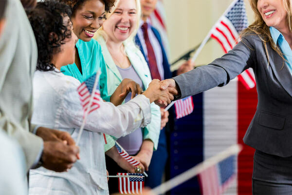 A smiling woman in a grey suit shakes hands with another woman in a white shirt. A diverse group of smiling people, many holding small American flags, are visible in the background.