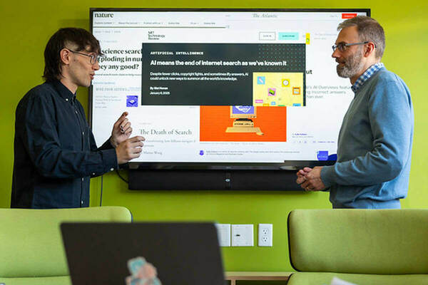 Two people discuss articles displayed on a large monitor featuring headlines about the impact of AI on internet searches. The person gesturing toward the screen wears a dark shirt. The other, wearing a blue sweater, listens attentively. Articles from MIT Technology Review, Nature, and The Atlantic are visible.