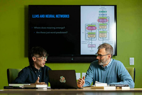 Two people converse at a table with a laptop and books. A monitor on the lime green wall displays a presentation slide titled "LLMs and Neural Networks." The slide asks, "Where does meaning emerge?" and "Are these just word-predictors?", alongside a diagram.