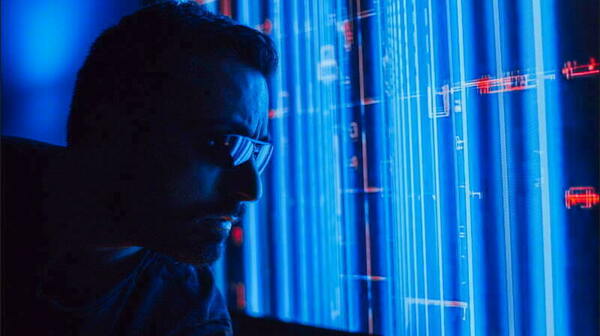 Person wearing glasses intently examines a computer screen displaying glowing blue vertical lines of code with small red accents.