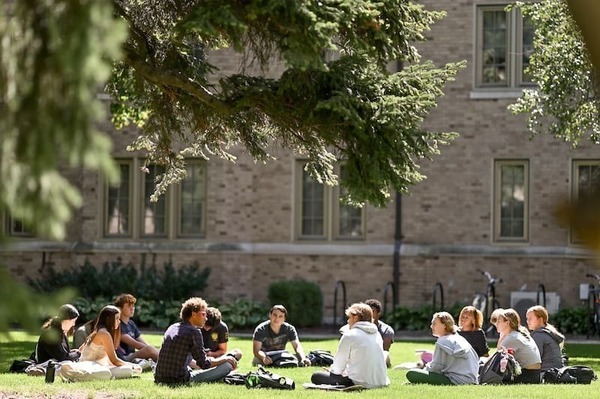 Students sit in a circle on the grass beneath a tree, engaged in discussion, in front of a University of Notre Dame building.