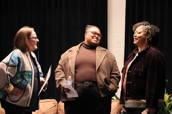 Three people smile and converse on a stage. One in a patterned quilted jacket, one in a brown plaid blazer, one in a plum jacket.