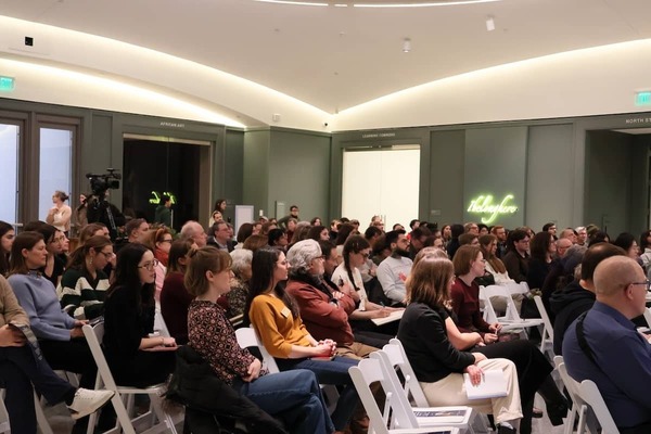 Audience members sit in white chairs at the Raclin Murphy Museum of Art, under a neon "I belong here" sign.