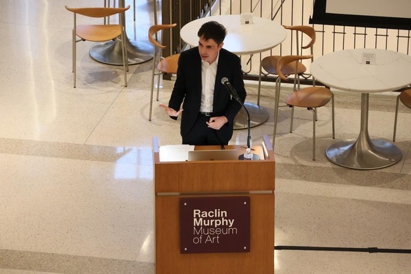 Young man in dark suit speaks, gesturing with hands, at a podium in the Raclin Murphy Museum of Art.