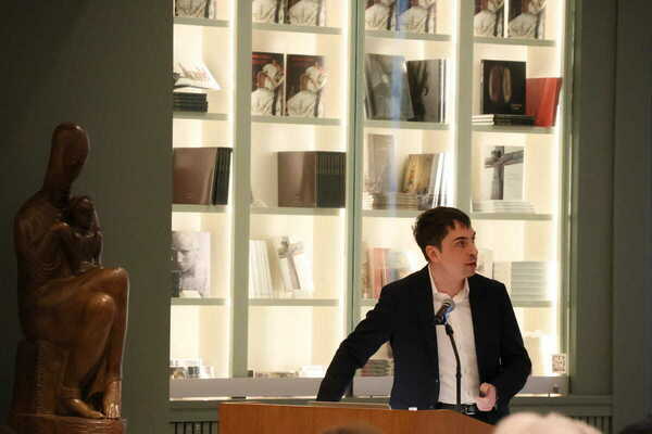 A man in a dark jacket speaks into a microphone at a podium. Behind him is a seated wooden statue and illuminated bookshelves.