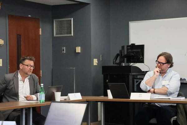 Two men sit at a table. One in a grey suit speaks while the other in a blue shirt listens thoughtfully, hand on chin.